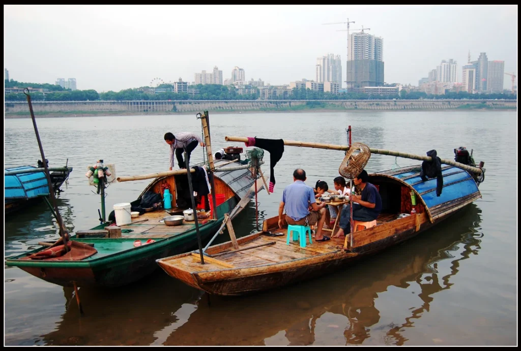 Several fishing boats docked at the riverbank, with people working and a city skyline in the background.